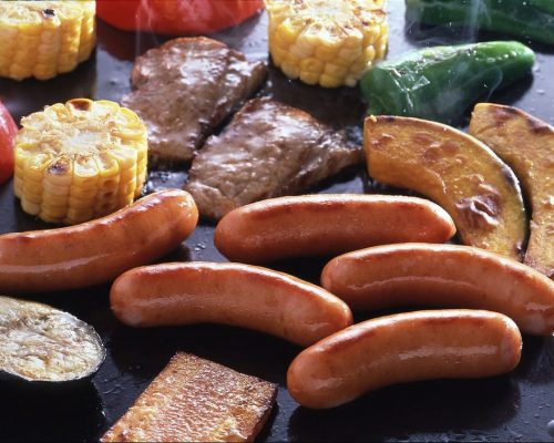 NH Foods Japanese Arabiki pork sausages grilling on a flat top BBQ with corn on the cob, kabocha squash, and sliced beef.